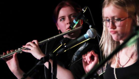 To jenter på scenen. En spiller fløyte, den andre står foran en mikrofon. 