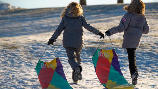 To unge jenter som løper på snøen med en fargerik akematte hver i hånden.
