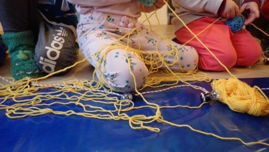 Et barn leker på gulvet med lange gule tråder fra et garnnøste.