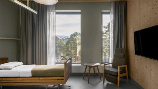 BIlde av pasient rom som viser seng, store vinduer, bord, stol og et stort TV som henger på vegg. Bilde er tatt av Fotograf : Hugo Lütcherath.