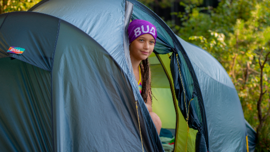 Grønt telt. Jente med lilla lue stikker hodet ut av teltet. Det står BUA på den lilla luen hennes.