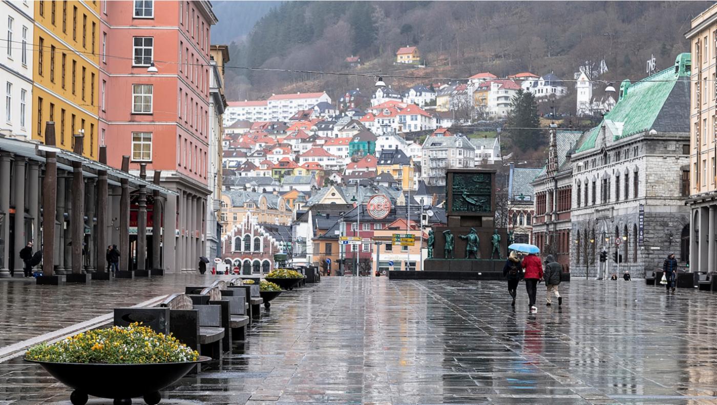 Torgalmenningen på en regnfull dag.Blomsterurner og bygninger venstre, byen oppover mot Skansen og fjellet rett frem og tre personer med paraply som spaserer mot Sjøfartmonumentet