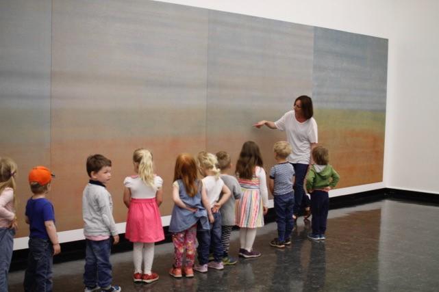 En rekke med barn ser på et kunstverk mens en voksen dame peker og forklarer. 