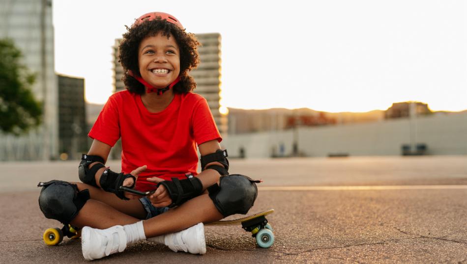 Et barn sitter på et skateboard og smiler med en telefon i hånden. 