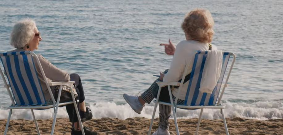To gråhårete damer i hver sin blå og hvitstripede solstol på stranden prater og ser ut mot det blå havet