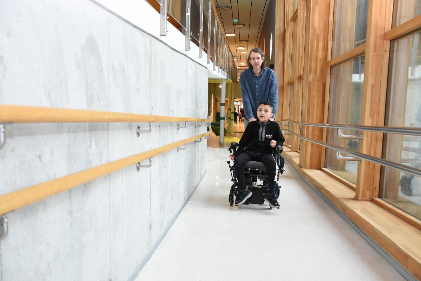 Person triller gutt i rullestol i korridor