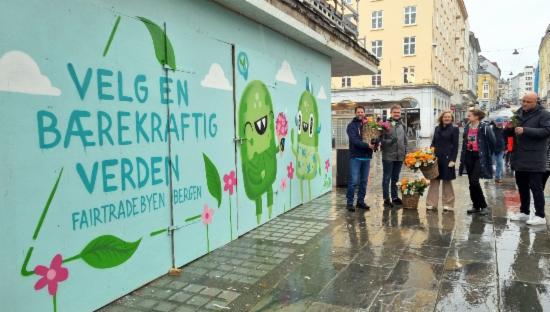 En gruppe mennesker med blomster står ved gatekunst med to figurer som gir blomster til hverandre. Blomstene er merket "Fairtrade". På veggen står "Velg en bærekraftig verden".