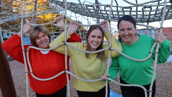 Tre frivillighetskoordinatorer i fargerike gensere i et klatretau på en lekeplass. 