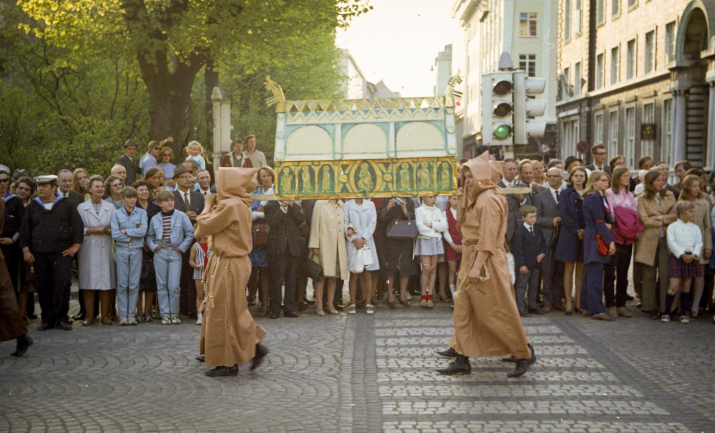 To personer utkledd som munker bærer på installasjon i et opptog. Folk står og ser på.