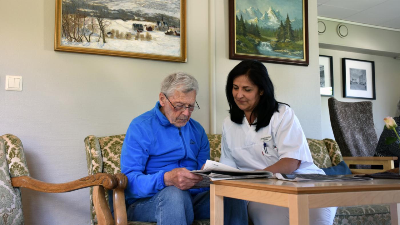 Sykepleier og beboer på sykehjem som sitter ved et bord og leser avisen. 