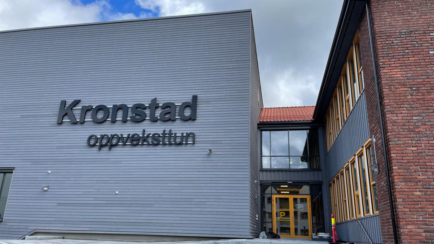 Stort grått bygg med bokstavane Kronstad oppveksttun på, knytt saman med bygning i raud teglstein.