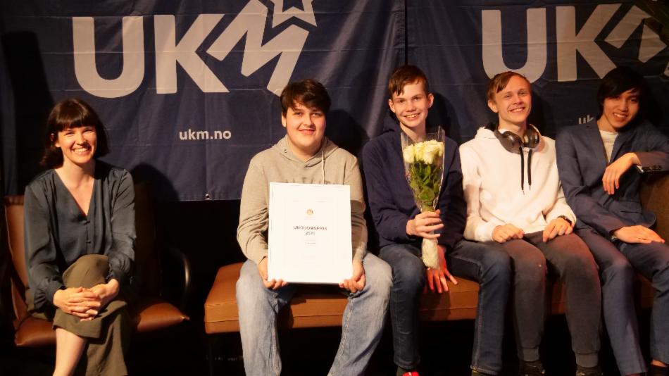 Bildet viser byråden sittende på en benk sammen med de fire ungdommene fra Knutepunkt. En av ungdommene holder et innrammet diplom, en annen en blomsterbukett. Alle smiler.  