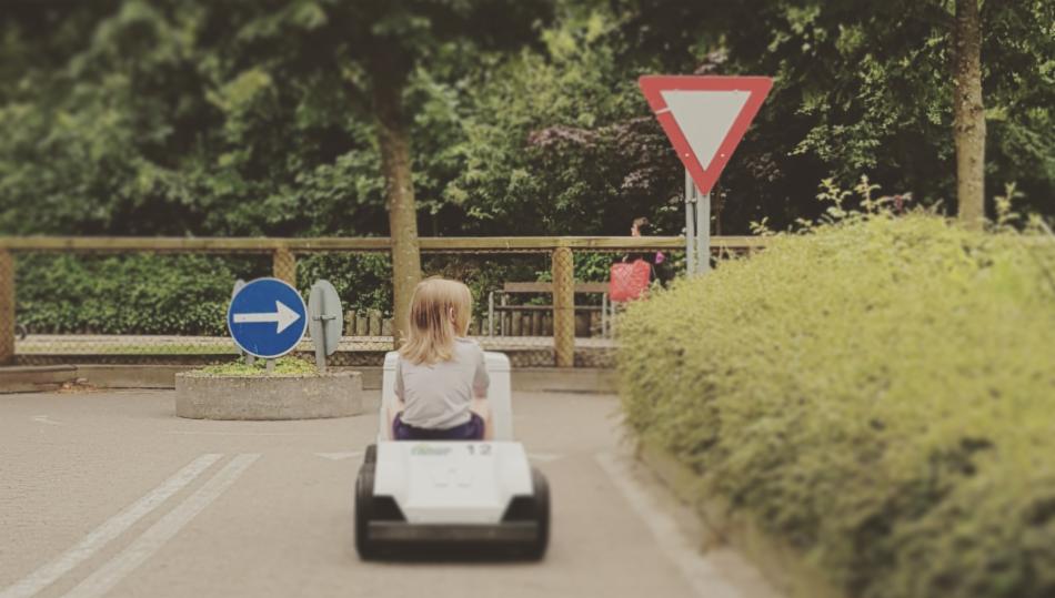 Et barn kjører liten bil på bane, med synlige trafikkskilt. 