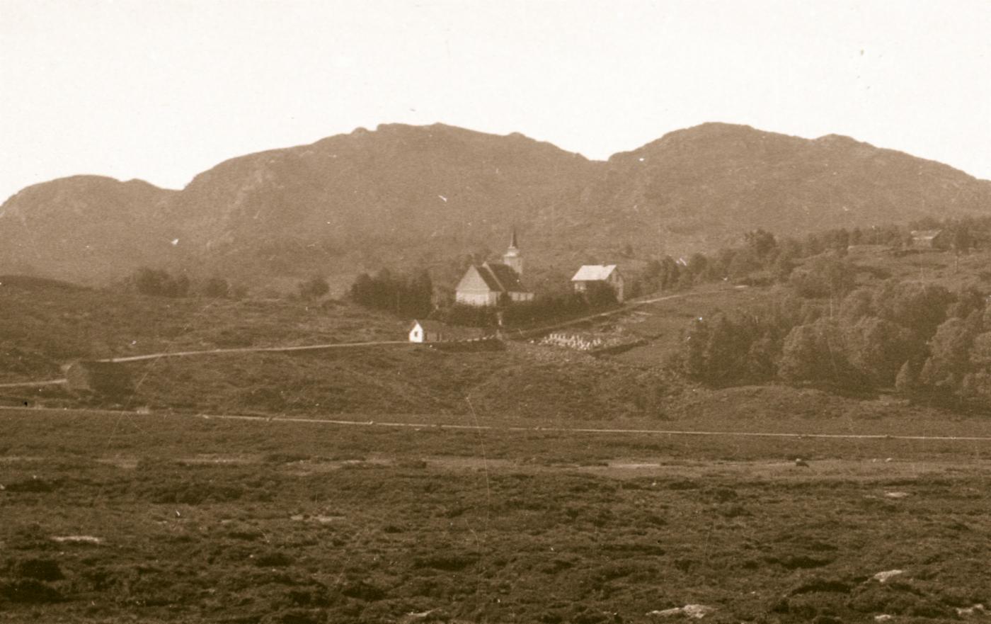 Det første kommunehuset i Åsane lå opprinnelig like ved kirken, slik dette fotografiet viser. I 1935 ble det flyttet nærmere kirkegården, der bårehuset står i dag. Huset ble revet i 1962. Utsnitt av foto fra arkivet etter Stadsingeniøren i Bergen, BBA-0193. Ukjent fotograf.